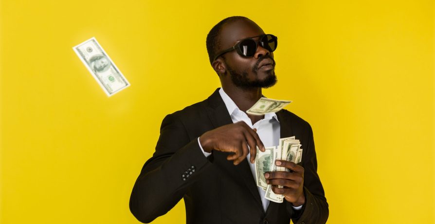 bearded serious young afroamerican guy is throwing out dollars from one hand, wearing sunglasses and black suit on the yellow background